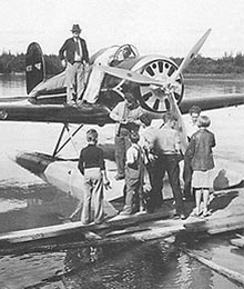 Photo of the Wiley Post-Will Rogers airplane refueling on the Chena River. Will Rogers and Wiley Post are signing autographs for the Spencer family children gathered on the dock. August 15, 1935
